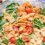 A large glass bowl with chicken and pasta with spinach, tomatoes and a hummus sauce and a text box on the photo