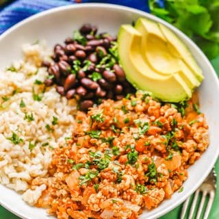 Low white bowl with chicken picadillo topped with cilantro served with rice, black beans and avocado