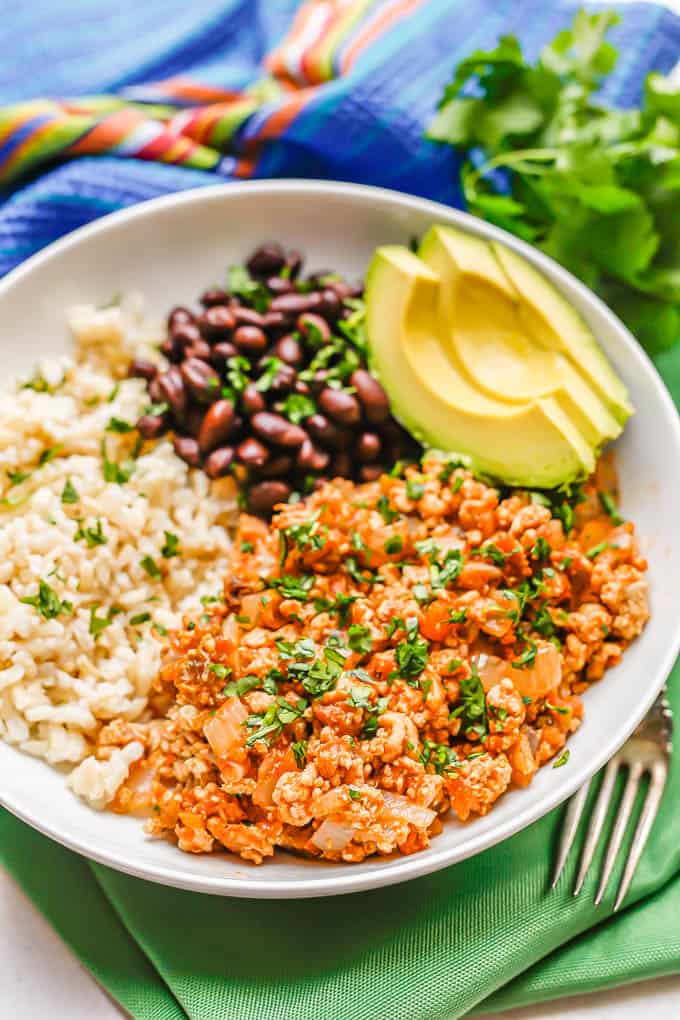 Low white bowl with chicken picadillo topped with cilantro served with rice, black beans and avocado