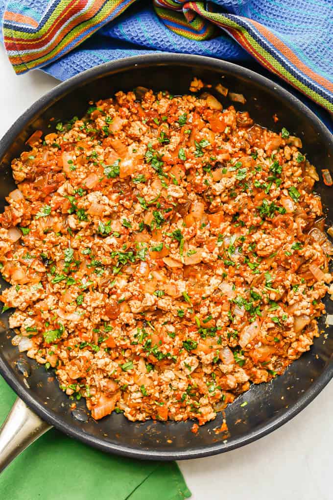 Cooked chicken picadillo in a large dark skillet with cilantro on top and colorful napkins to the side