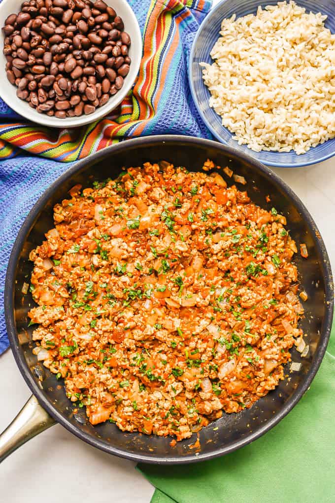 A large skillet with a ground chicken mixture topped with cilantro and bowls nearby of steamed rice and black beans