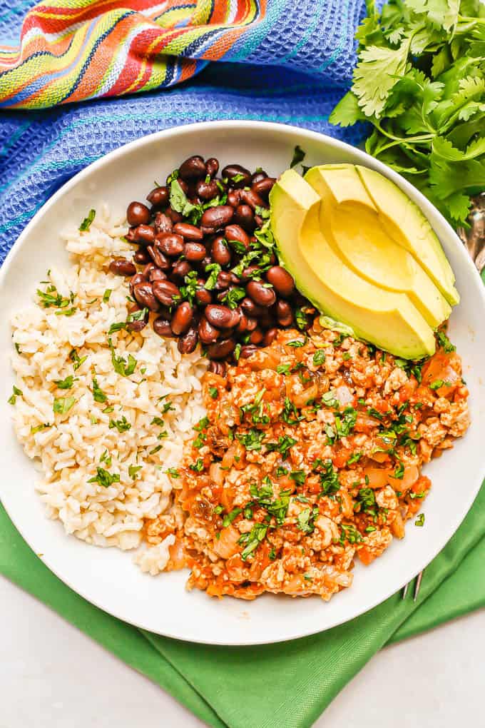 Chicken picadillo served in a low white bowl with rice, black beans and avocado with cilantro sprinkled on top and sprigs to the side