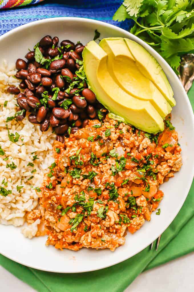 Chicken picadillo served in a low white bowl with rice, black beans and avocado with cilantro sprinkled on top