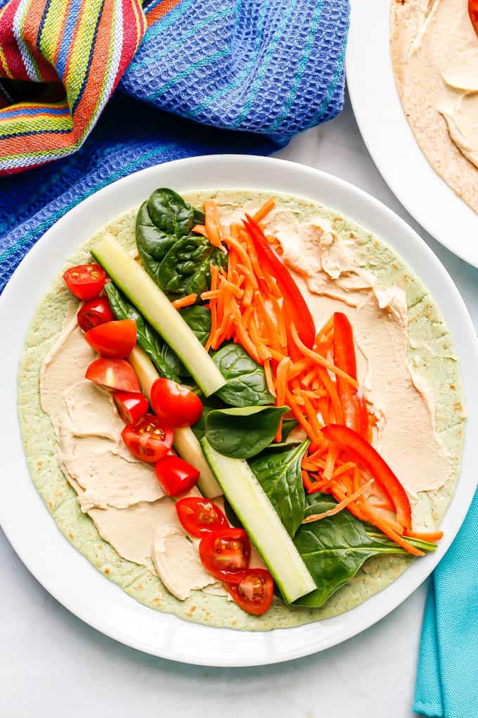 A tortilla spread with hummus and topped with colorful fresh veggies with colorful napkins nearby