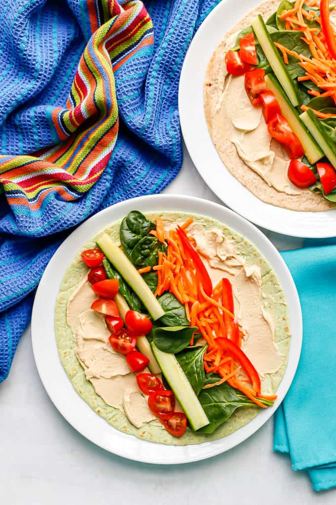 Two white plates with spinach tortillas spread with hummus and topped with colorful veggies
