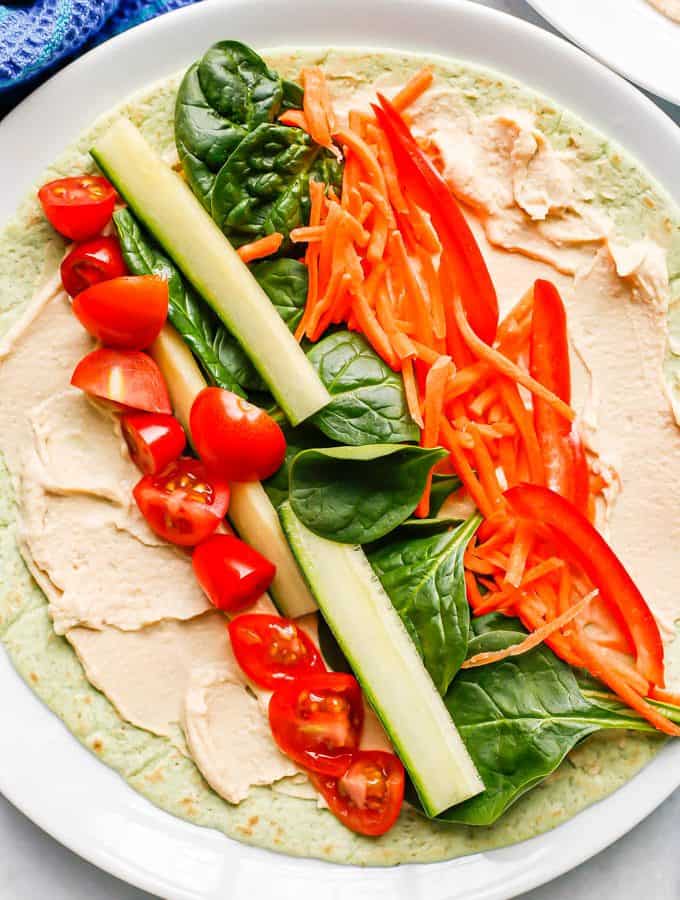 A white plate with a spinach tortilla layered with hummus, tomato, cucumber, spinach, carrots and peppers