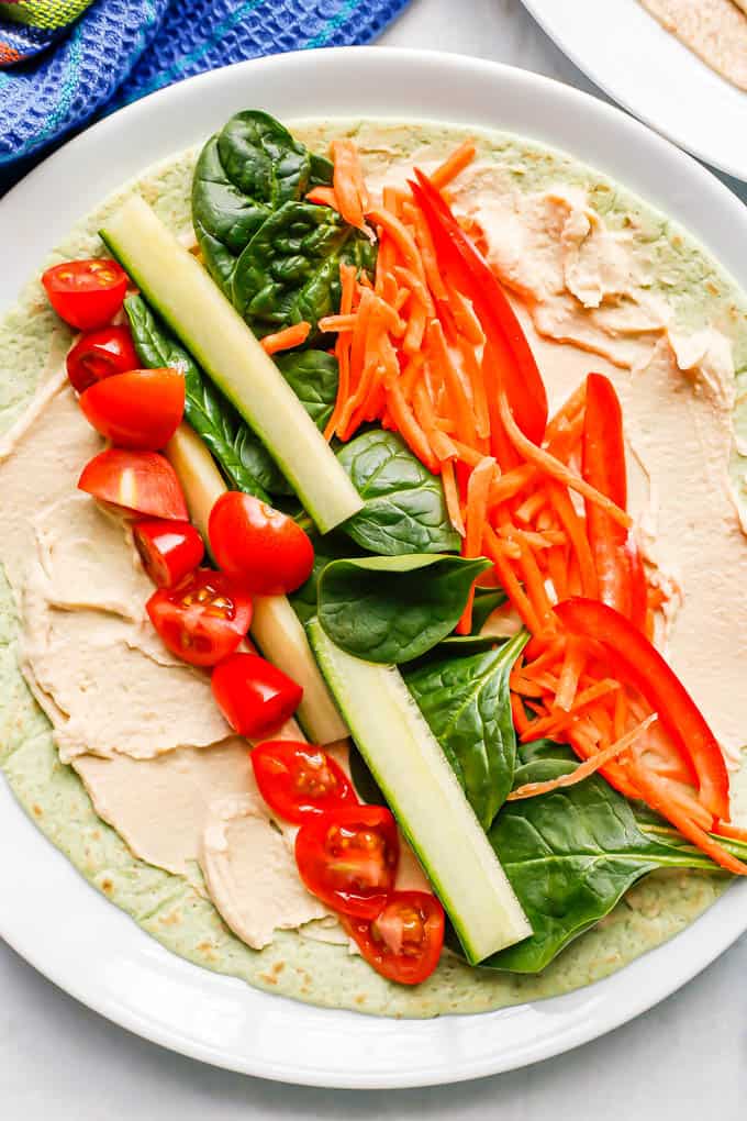 A white plate with a spinach tortilla layered with hummus, tomato, cucumber, spinach, carrots and peppers