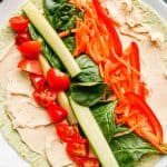 A white plate with a spinach tortilla layered with hummus, tomato, cucumber, spinach, carrots and peppers with a text box on the photo