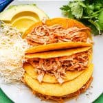 Shredded chicken tacos served on a plate with cheese, avocado and cilantro and a text overlay on the photo