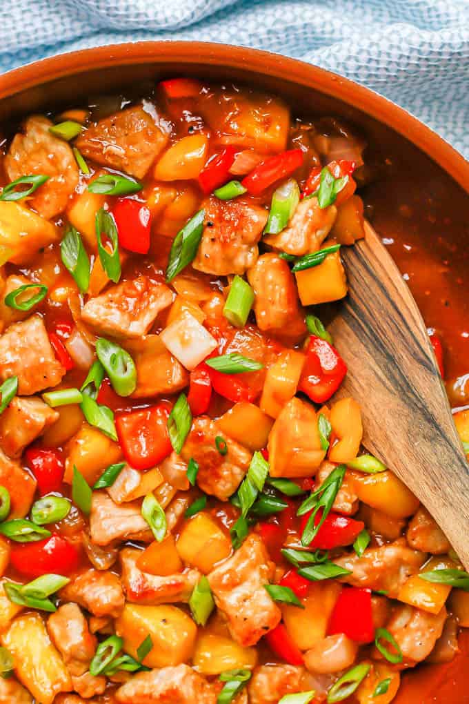 A wooden spoon in a large copper skillet of sweet and sour pork with peppers and pineapple