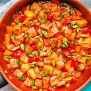 Sweet and sour pork with peppers and pineapple in a large copper skillet with sliced green onions on top and teal napkins nearby