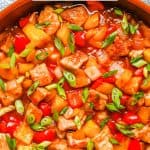 Sweet and sour pork with peppers and pineapple in a large copper skillet with sliced green onions on top and a text overlay on the photo