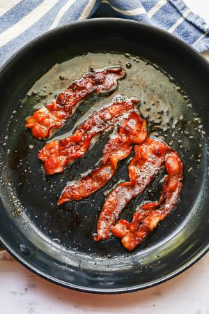 5 slices of bacon being cooked in a large dark skillet