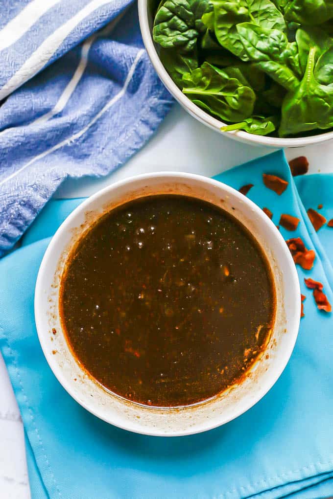 Warm bacon vinaigrette served in a white bowl with a bowl of spinach to the side and crumbled pieces of bacon scattered around