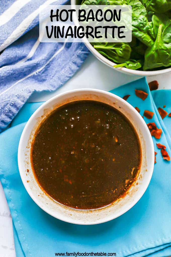 Warm bacon vinaigrette served in a white bowl with a bowl of spinach to the side and crumbled pieces of bacon scattered around and a text overlay on the photo