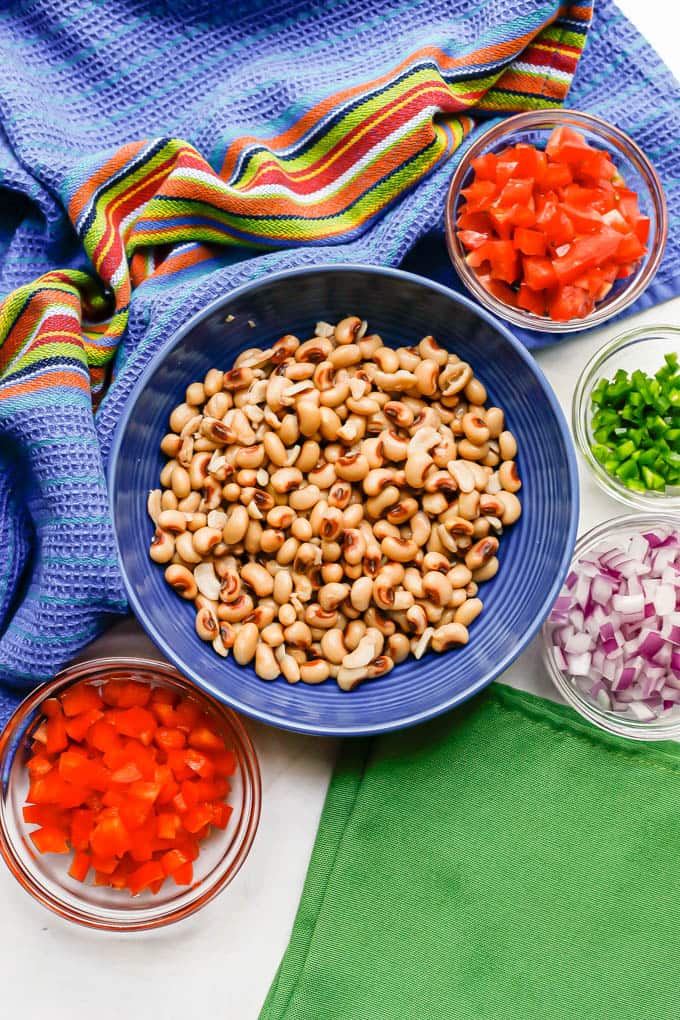 Separate bowls of peppers, tomatoes, black eyed peas, jalapeno and red onion laid out on a counter with colorful napkins