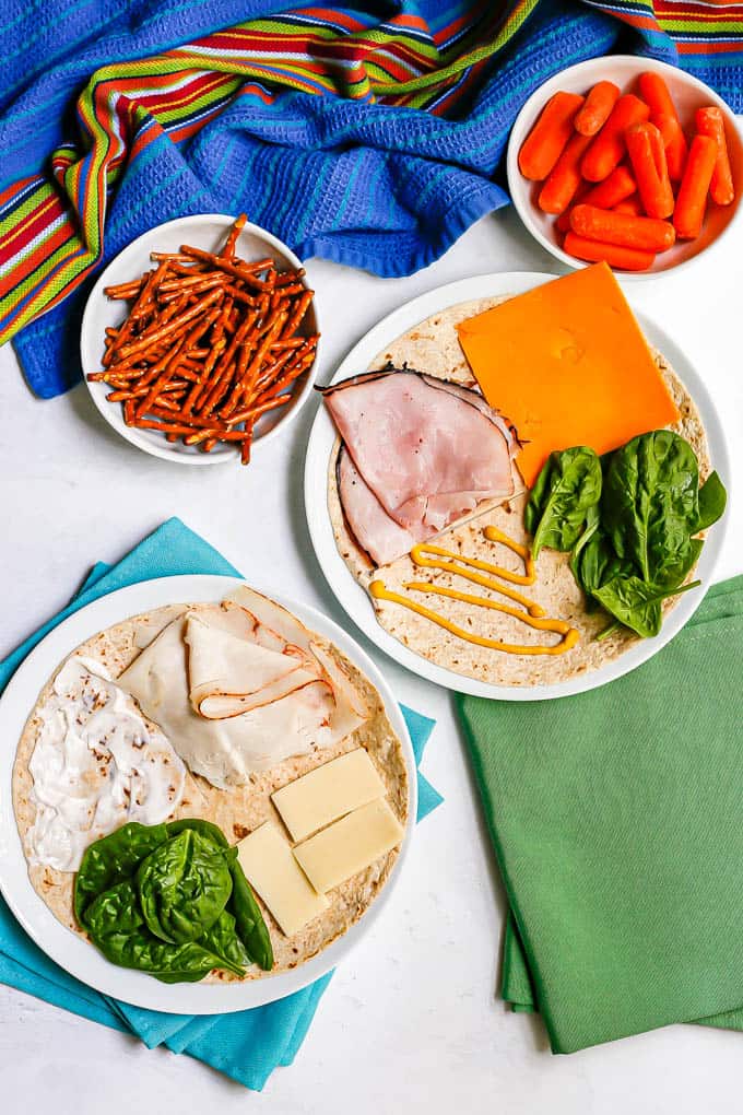 A ham and cheese and turkey and cheese tortilla on separate plates with spinach and spreads on tortilla before being folded