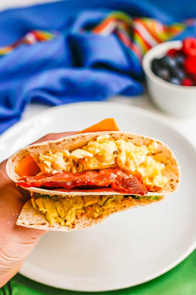 A hand holding up a folded breakfast wrap with scrambled eggs, bacon, cheese and avocado