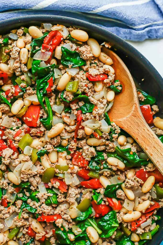 A wooden spoon resting in a large skillet with a ground turkey, white bean, tomato and spinach nmixture