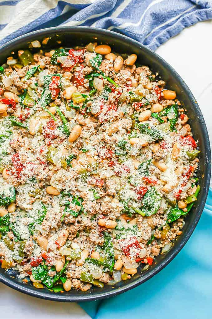 A large dark skillet with ground turkey, white beans, tomatoes and spinach with Parmesan cheese on top and colorful napkins to the side