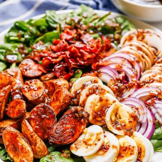 A spinach salad on a large white platter with roasted fingerling potatoes, sliced hard boiled eggs, red onion slices and sliced mushrooms, plus crumbled bacon and a bacon vinaigrette
