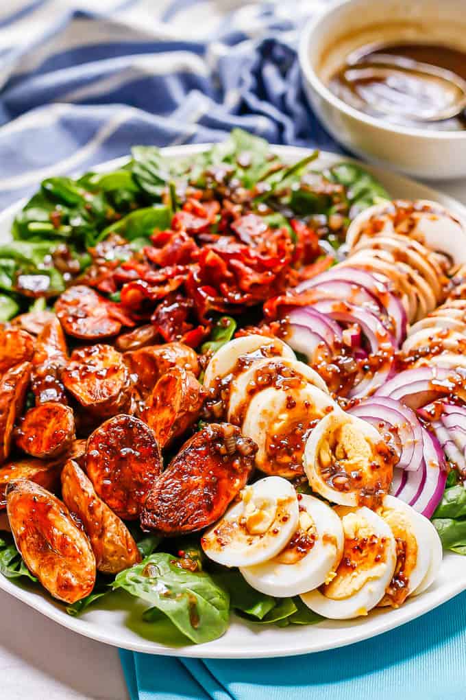 A spinach salad on a large white platter with roasted fingerling potatoes, sliced hard boiled eggs, red onion slices and sliced mushrooms, plus crumbled bacon and a bacon vinaigrette