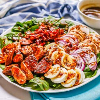 Horizontal image of a A spinach salad on a large white platter with roasted fingerling potatoes, sliced hard boiled eggs, red onion slices and sliced mushrooms, plus crumbled bacon and a bacon vinaigrette