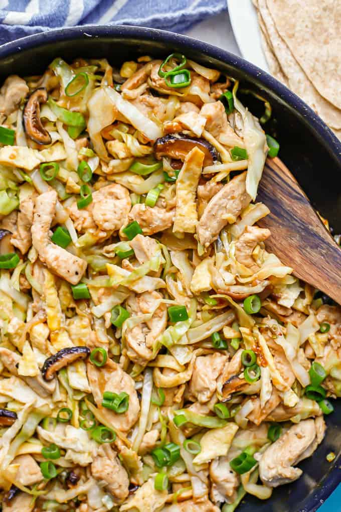 A wooden spoon resting in a skillet of homemade moo shu chicken with cabbage and mushrooms