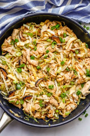 A large dark skillet with moo shu chicken with cabbage, mushrooms and egg topped with sliced green onions