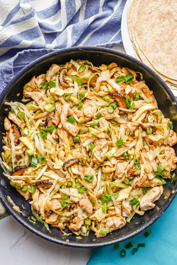 A large dark skillet with moo shu chicken topped with green onions and a white plate with tortillas to the side for serving