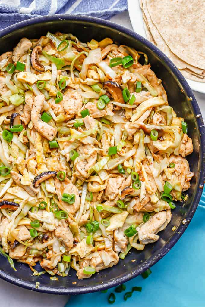 A large skillet with moo shu chicken with cabbage, mushrooms, egg and green onion on top with a plate of tortillas to the side