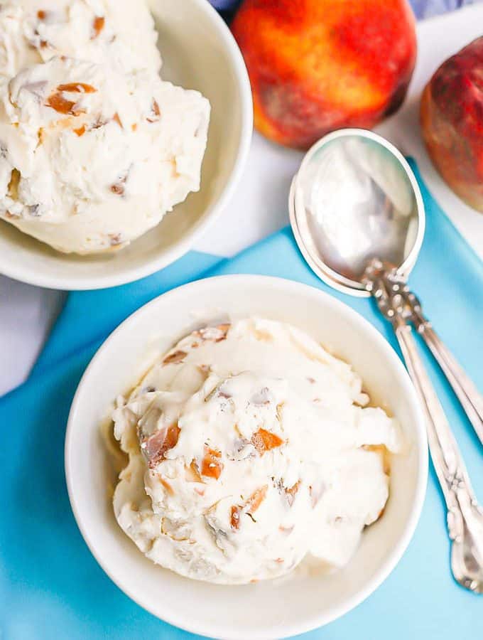 Two white bowls with scoops of homemade peach ice cream with some spoons and fresh peaches to the side