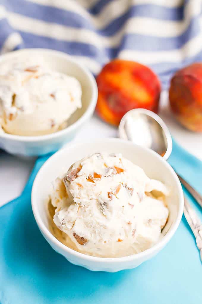 Scoops of homemade peach ice cream served in two white bowls with some spoons and fresh peaches to the side