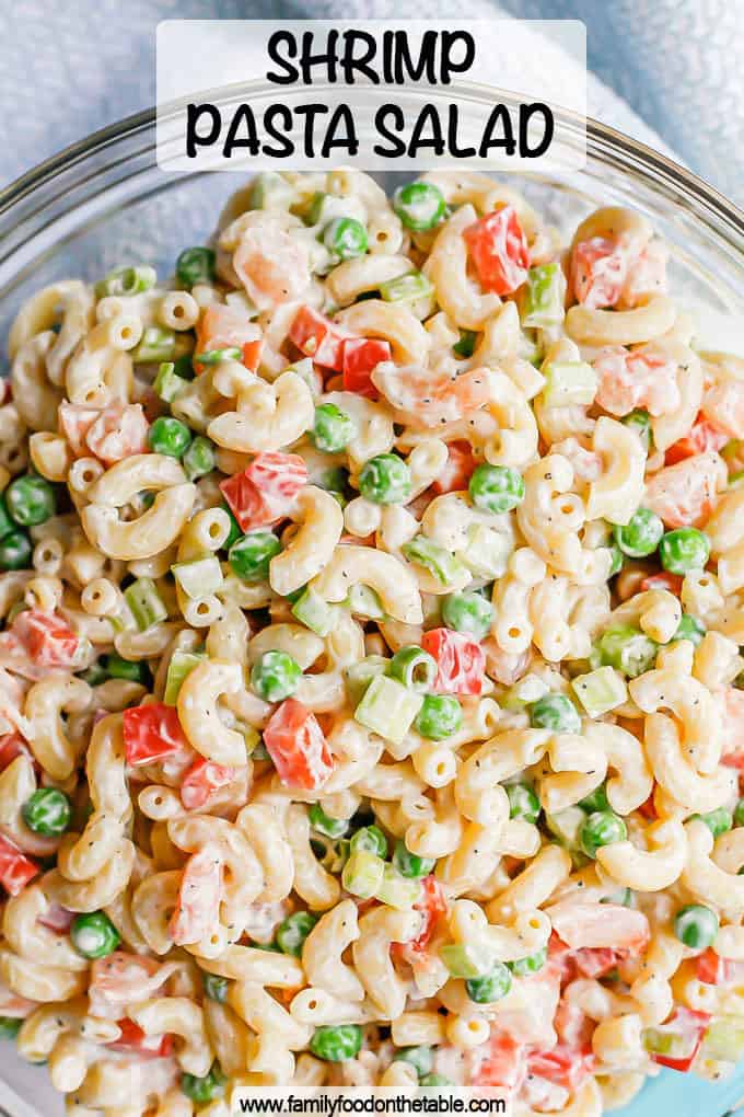 A cold, creamy shrimp pasta salad with peas and bell pepper in a large glass bowl with a text overlay on the image