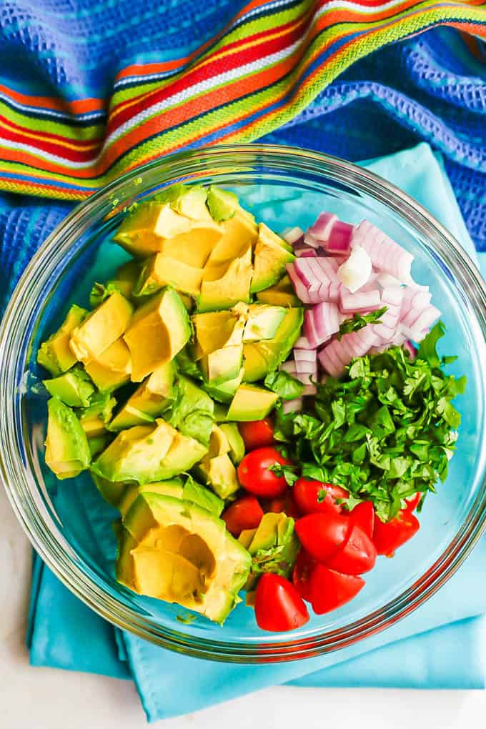 A clear glass bowl with layered ingredients of diced avocado, cherry tomatoes, red onion and chopped cilantro before being mixed together