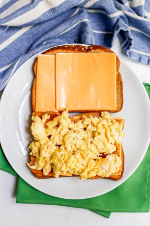 A breakfast sandwich being assembled with bread, American cheese, bacon and scrambled eggs