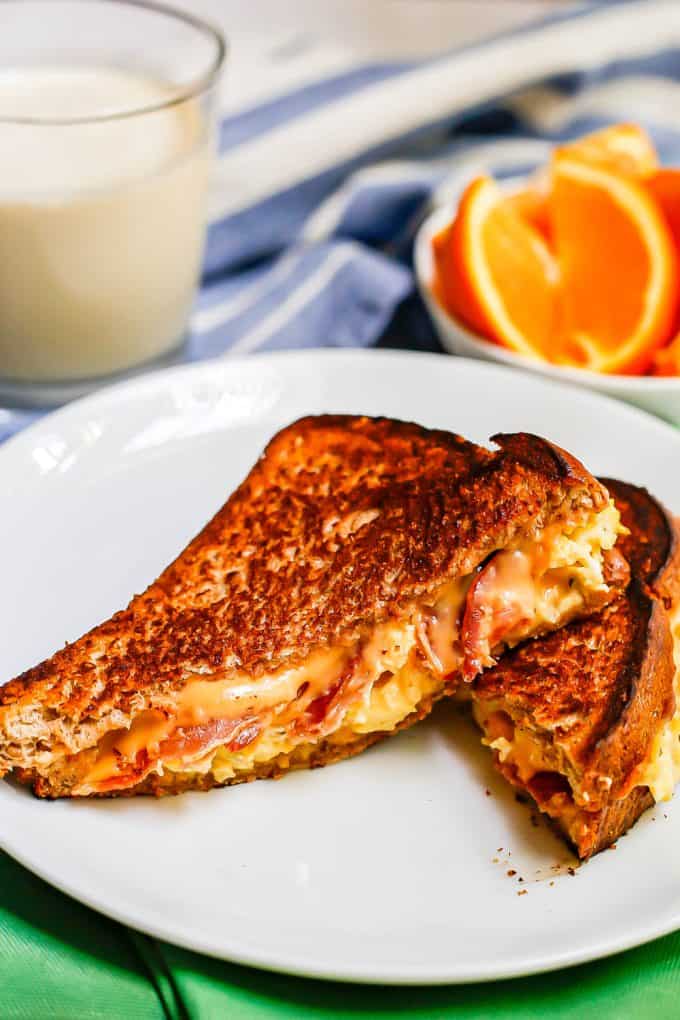 A grilled cheese with bacon and eggs cut in half and served on a white plate with milk and sliced oranges in the background