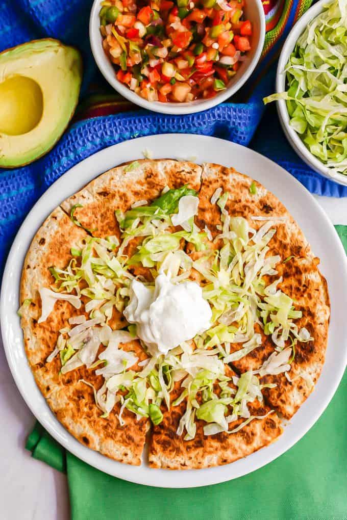 Chicken quesadilla pieces on a white plate with shredded lettuce and Greek yogurt on top and extra toppings in bowls nearby