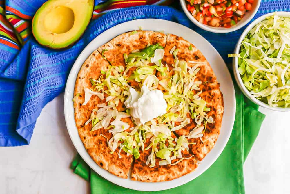 A whole chicken quesadilla, cut into pieces, on a white plate with shredded lettuce and Greek yogurt on top