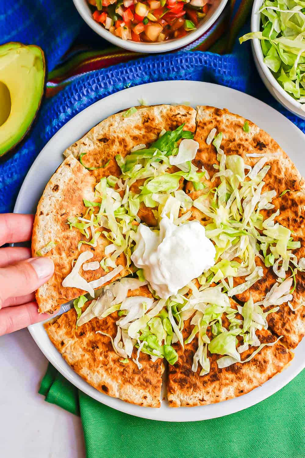 A hand picking up a wedge of chicken quesadilla topped with shredded lettuce and Greek yogurt from a plate