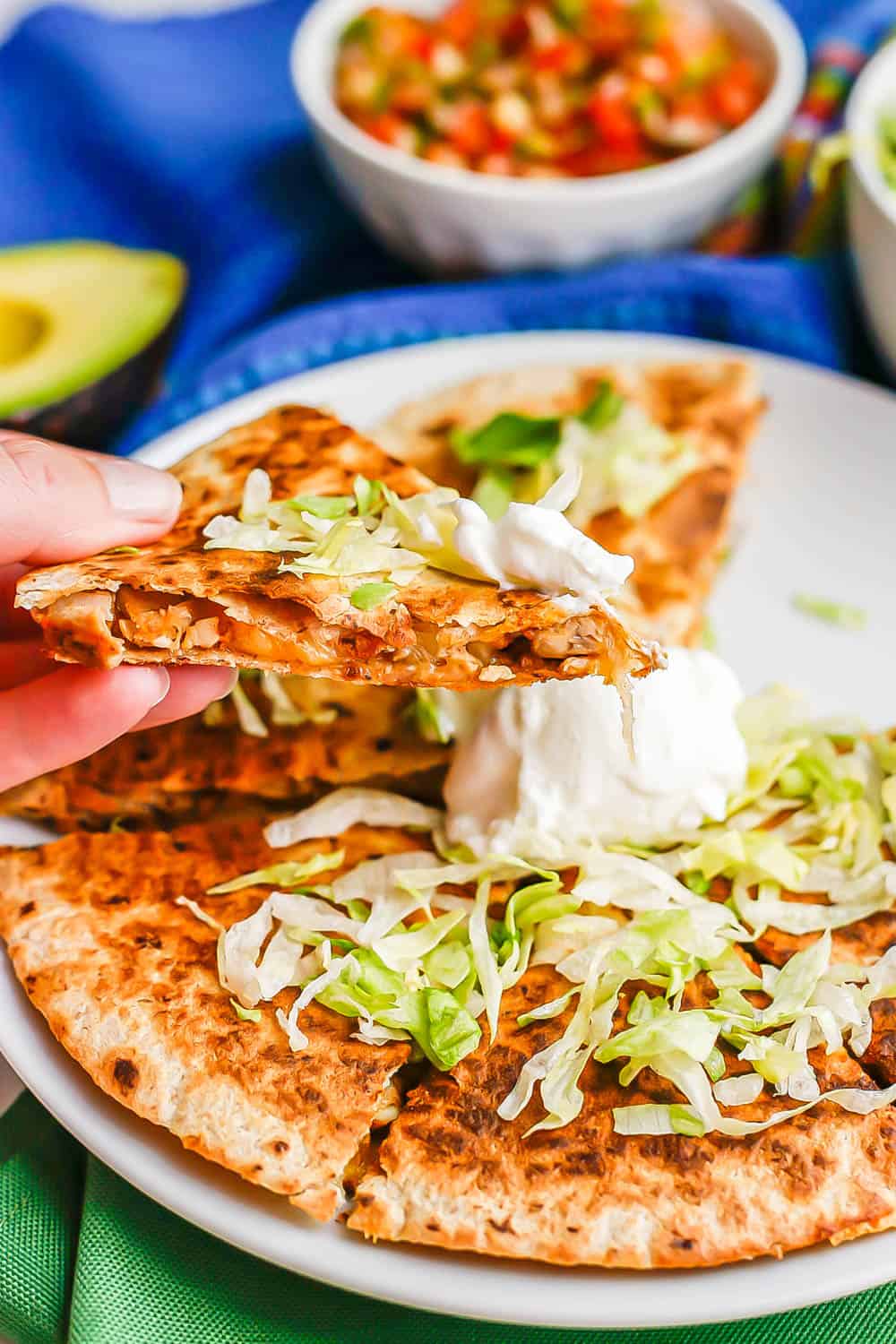 A hand holding up a cheesy chicken quesadilla piece with lettuce and Greek yogurt