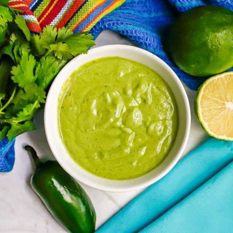 A creamy green cilantro lime dressing in a small white bowl with jalapeno, cilantro and limes nearby