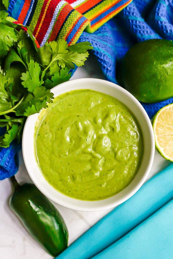 A creamy cilantro lime dressing in a small white bowl with jalapeno, cilantro and limes nearby