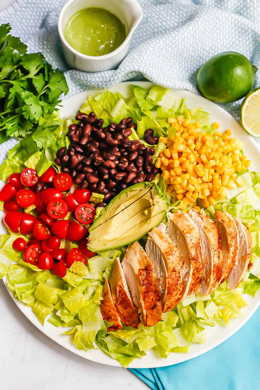 A white platter with romaine lettuce topped with arranged tomatoes, black beans, corn, avocado and sliced chicken breast with a white cruet filled with green salad dressing to the side