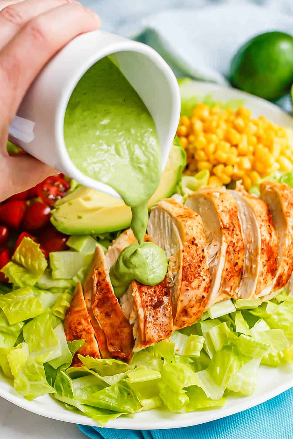 A green dressing being poured out of a white salad dressing cruet onto sliced chicken breasts on an arranged salad