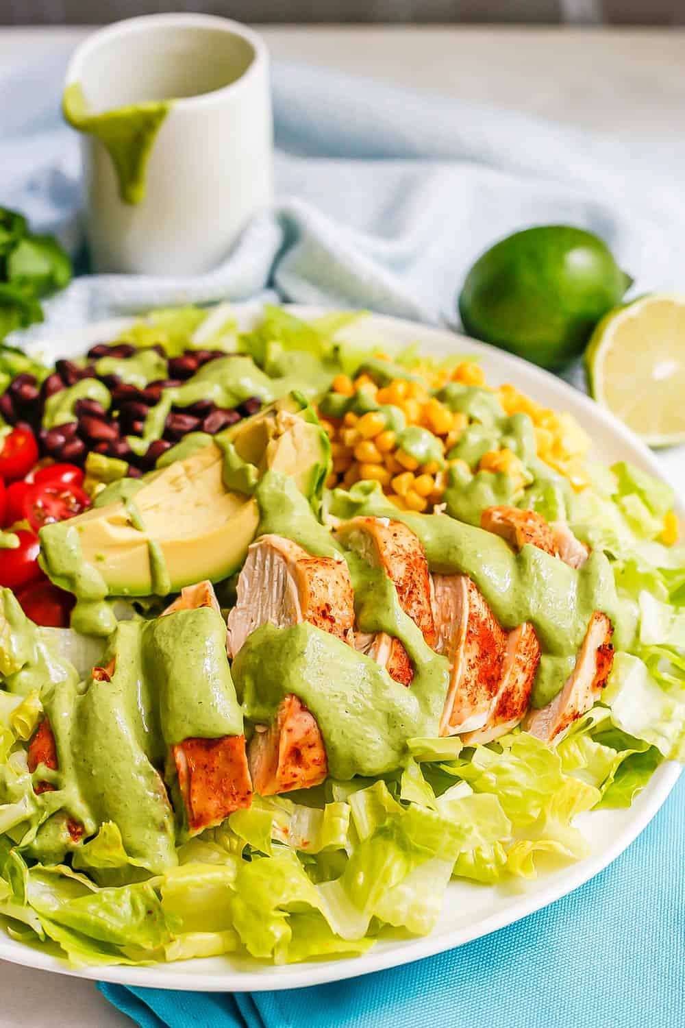 Close up of green cilantro lime dressing on sliced chicken breast on an arranged salad on a white platter with limes in the background