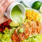 A hand holding a white cruet pouring a green cilantro lime dressing onto sliced chicken breast on a salad with a text overlay on the photo