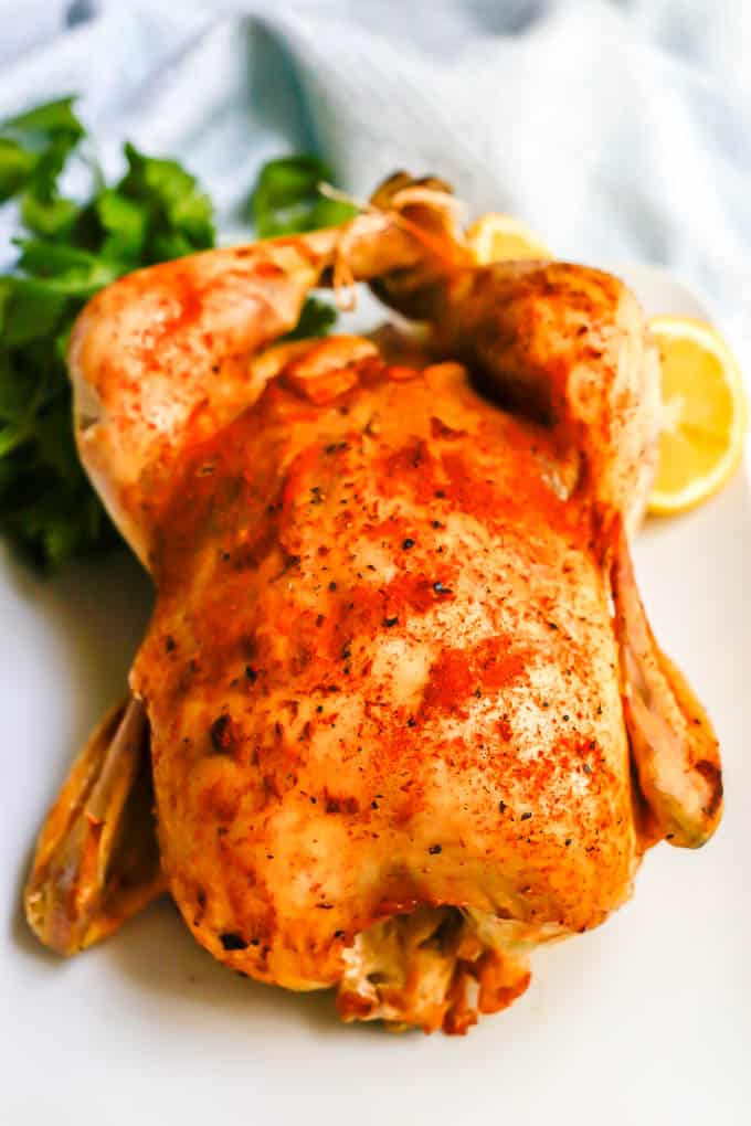 A seasoned and cooked whole chicken served on a white platter with lemon halves and fresh herbs in the background