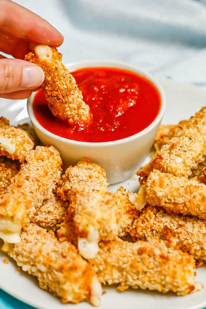 A hand dipping a breaded mozzarella stick into a small white bowl of marinara sauce