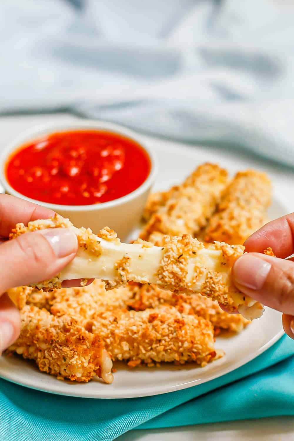 A hand pulling a cheesy breaded mozzarella stick apart in front of a plate of mozzarella sticks with marinara
